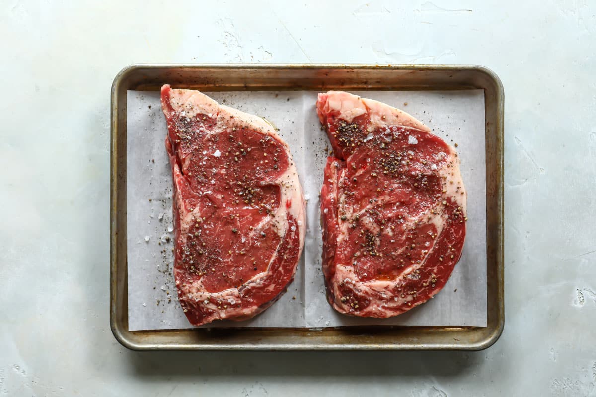 Two seasoned rib eyes on a baking sheet before being cooked.