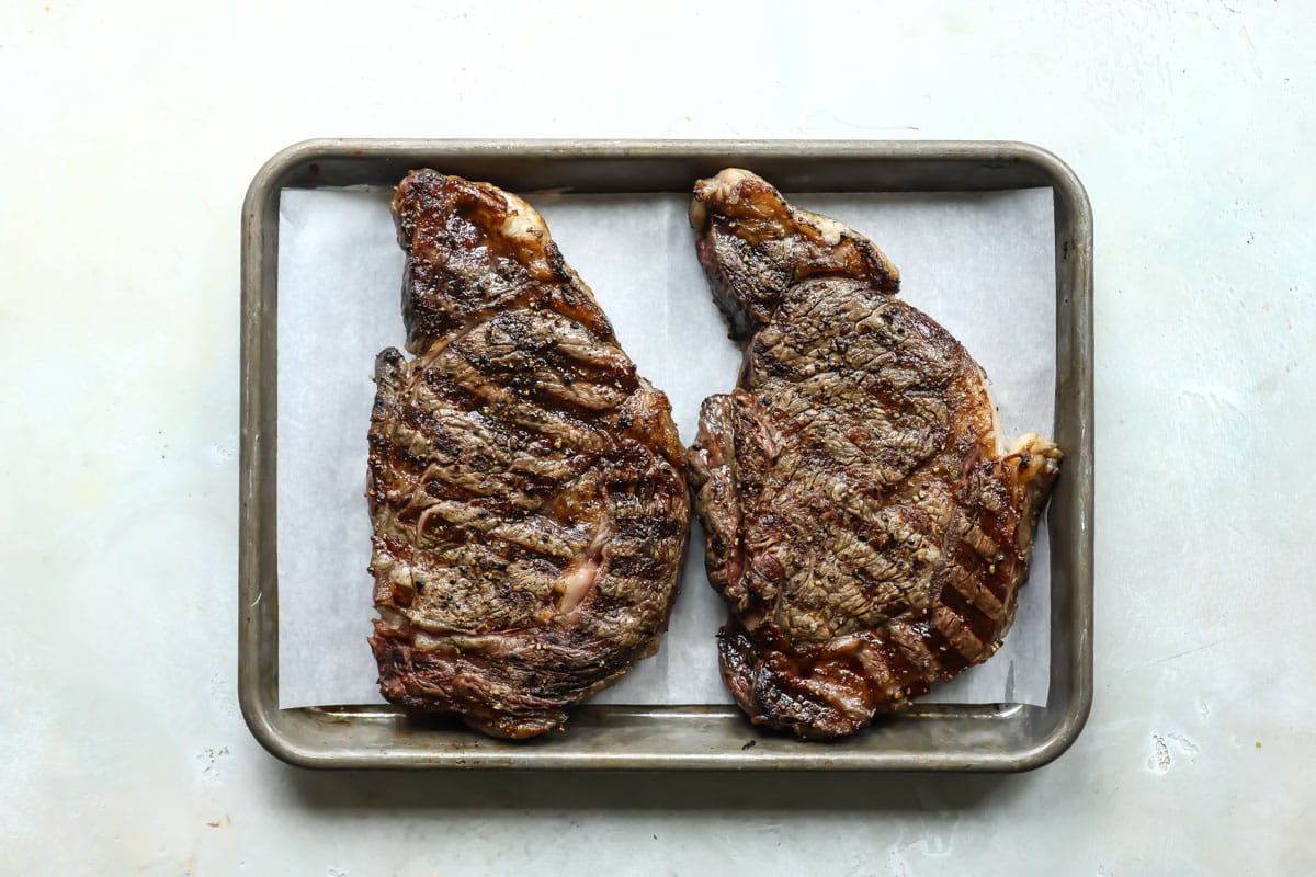 Two rib eyes on a parchment paper lined baking sheet.