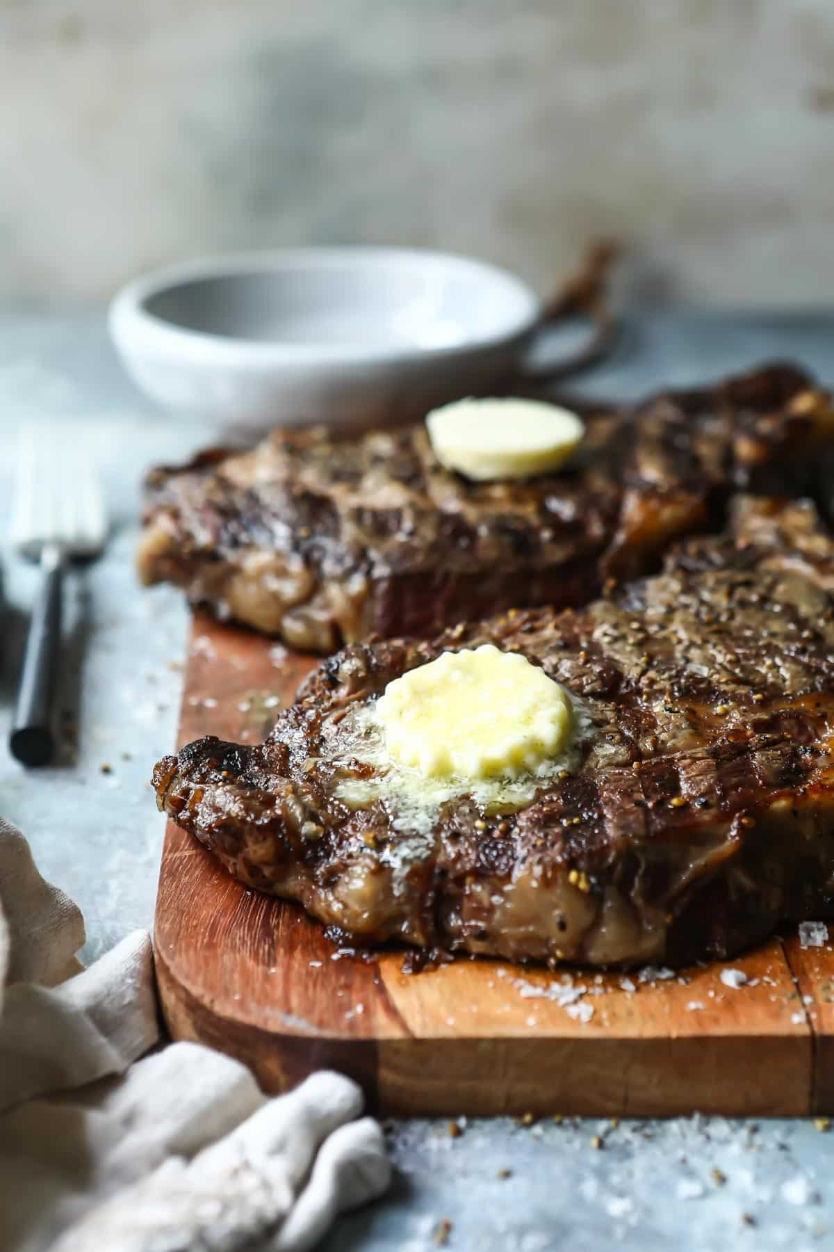 Two Rib Eye steaks on a wooden cutting board topped with butter.