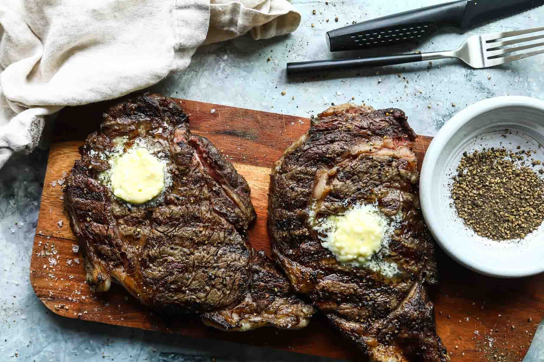 Two Rib Eye steaks on a wooden cutting board topped with butter.