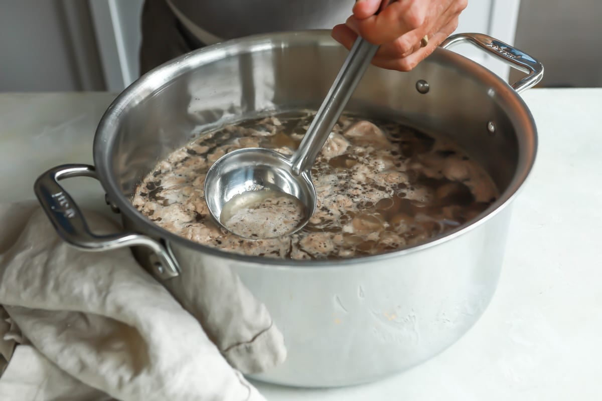 Fat being skimmed off of the top of a pot with water and a pork shoulder.