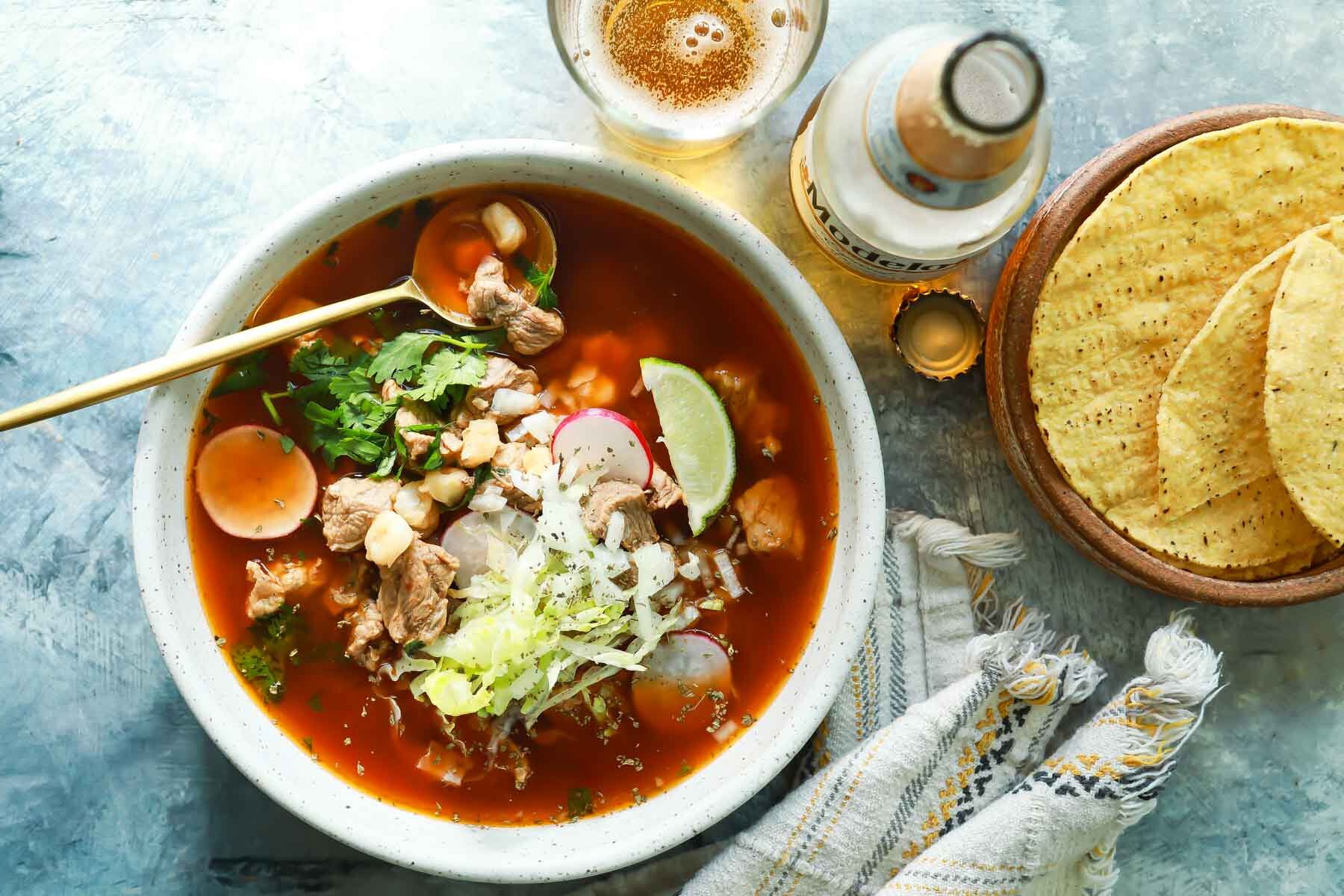 A bowl of pozole rojo next to a bottle of Modelo.