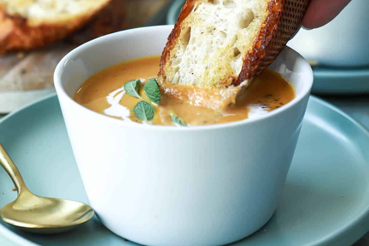 A piece of crusty bread being dipped in pumpkin soup.