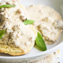A white bowl of biscuits smothered in sausage gravy garnished with sage leaves.