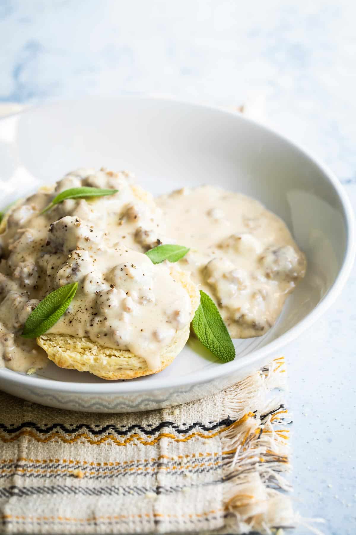 A white bowl of biscuits smothered in sausage gravy garnished with sage leaves.