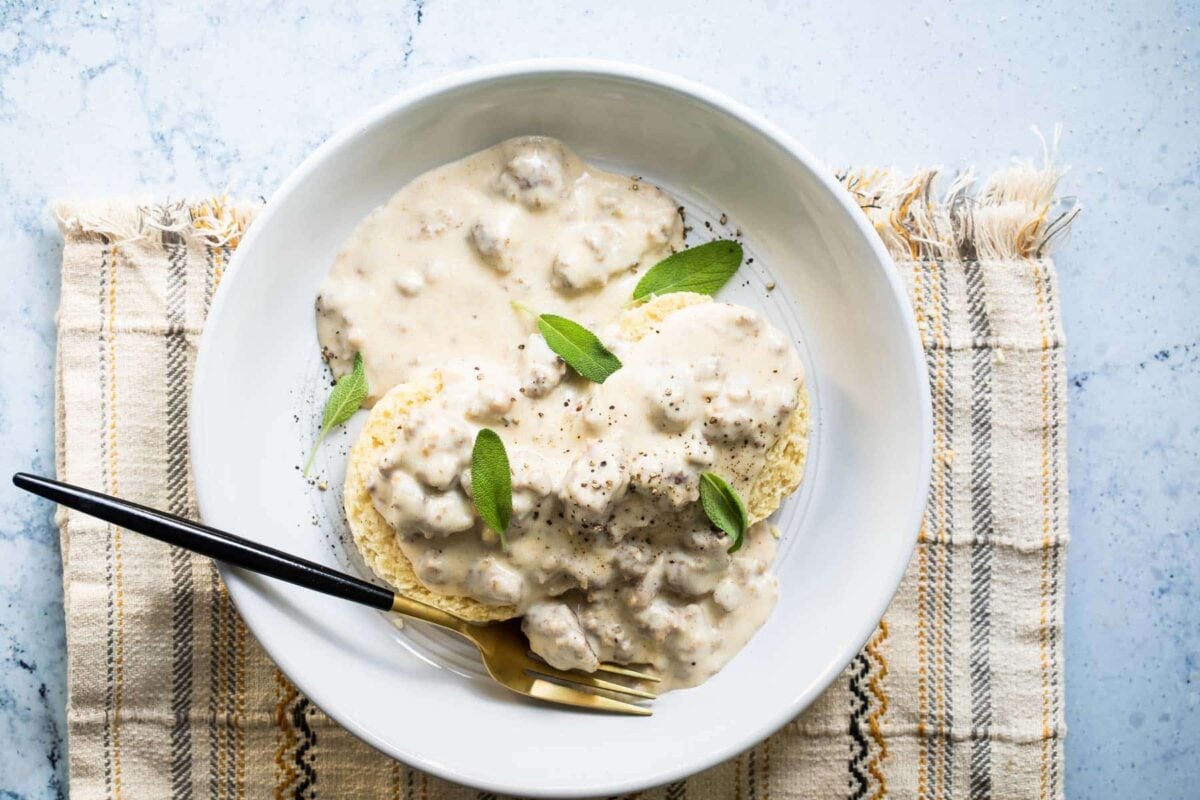 A white bowl of biscuits smothered in sausage gravy garnished with sage leaves.