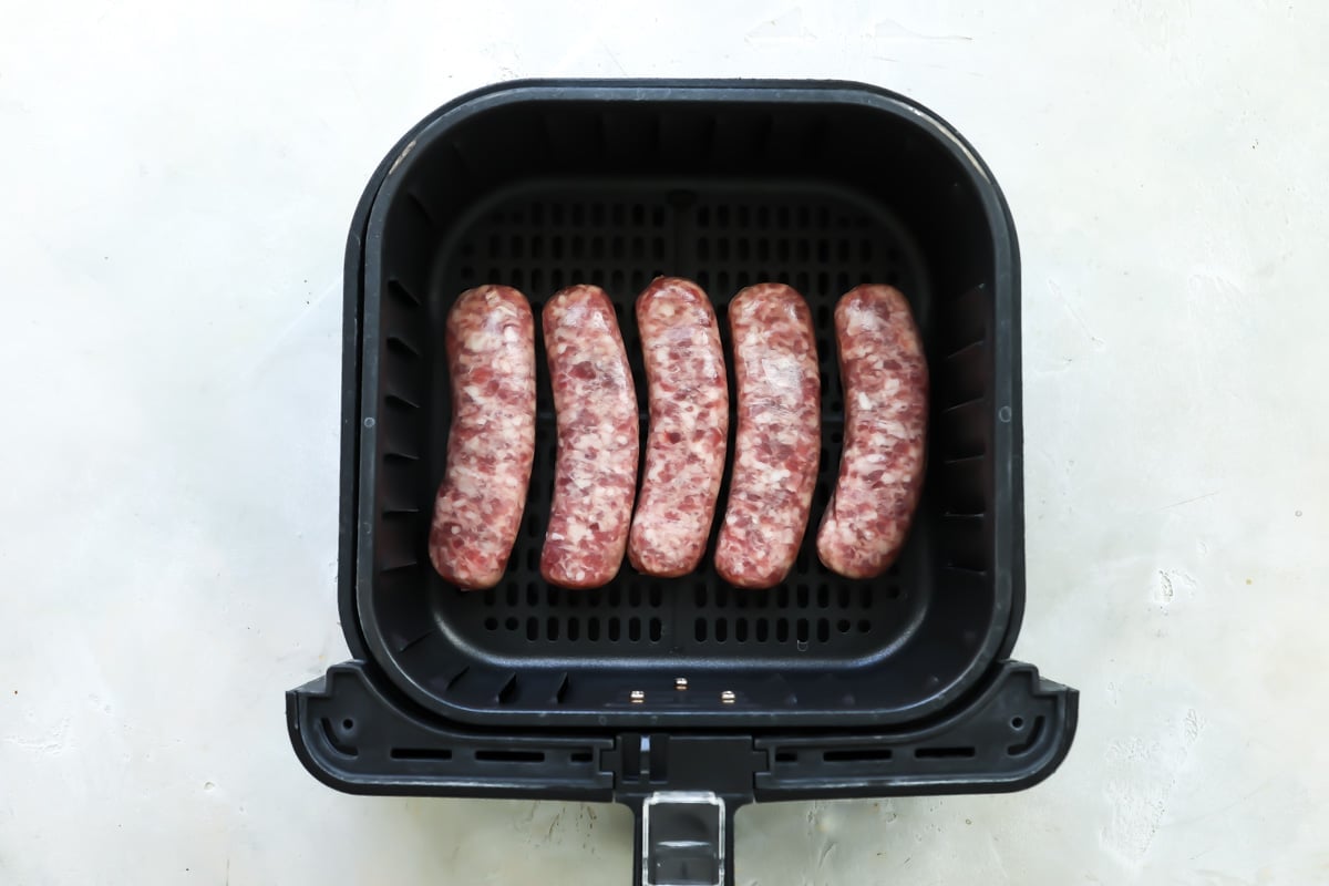An air fryer basket with uncooked bratwursts in it.