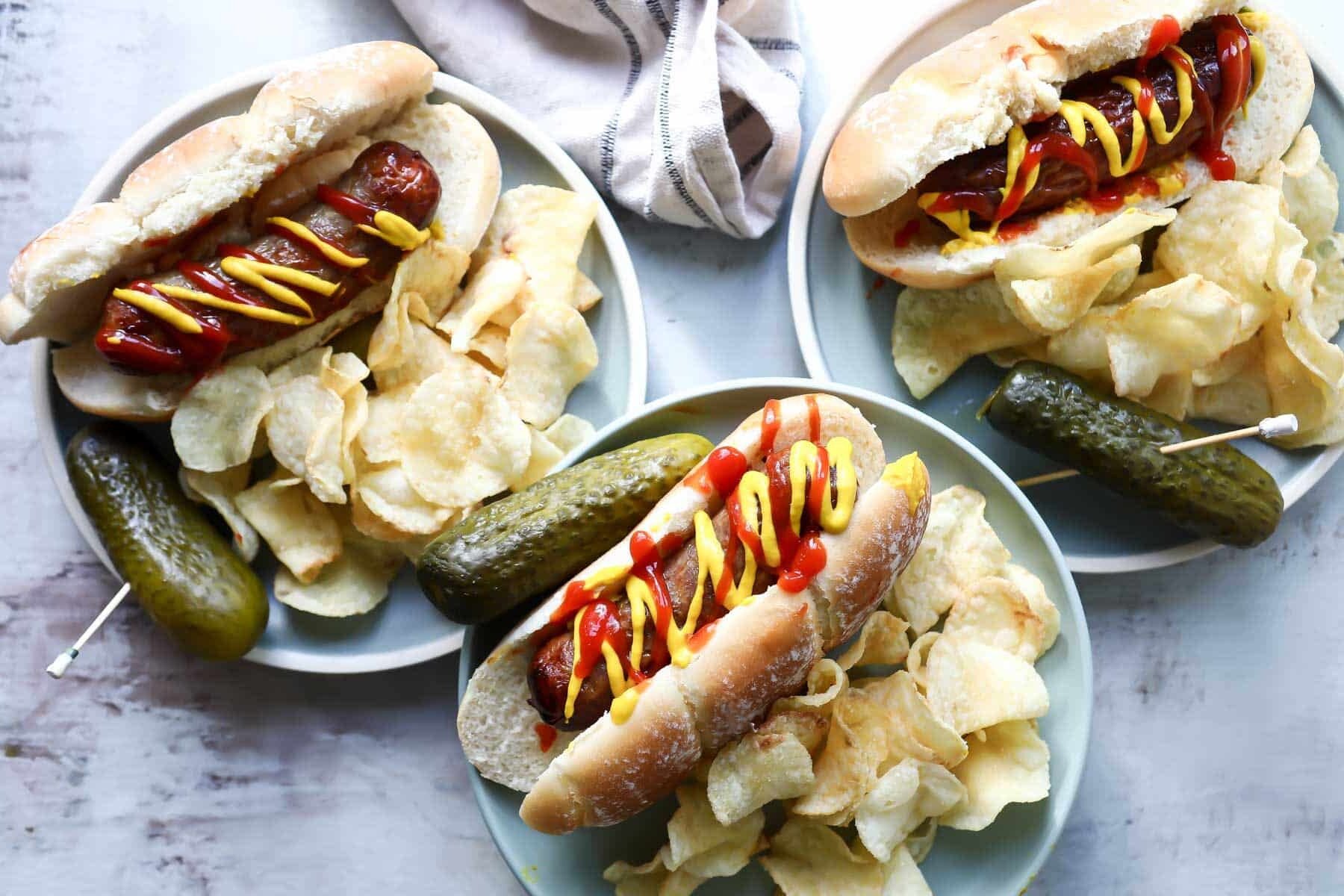 Three plates with air fryer bratwurst on plates with potato chips and a pickles.
