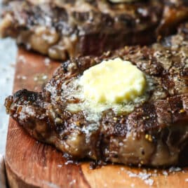 Two Rib Eye steaks on a wooden cutting board topped with butter.