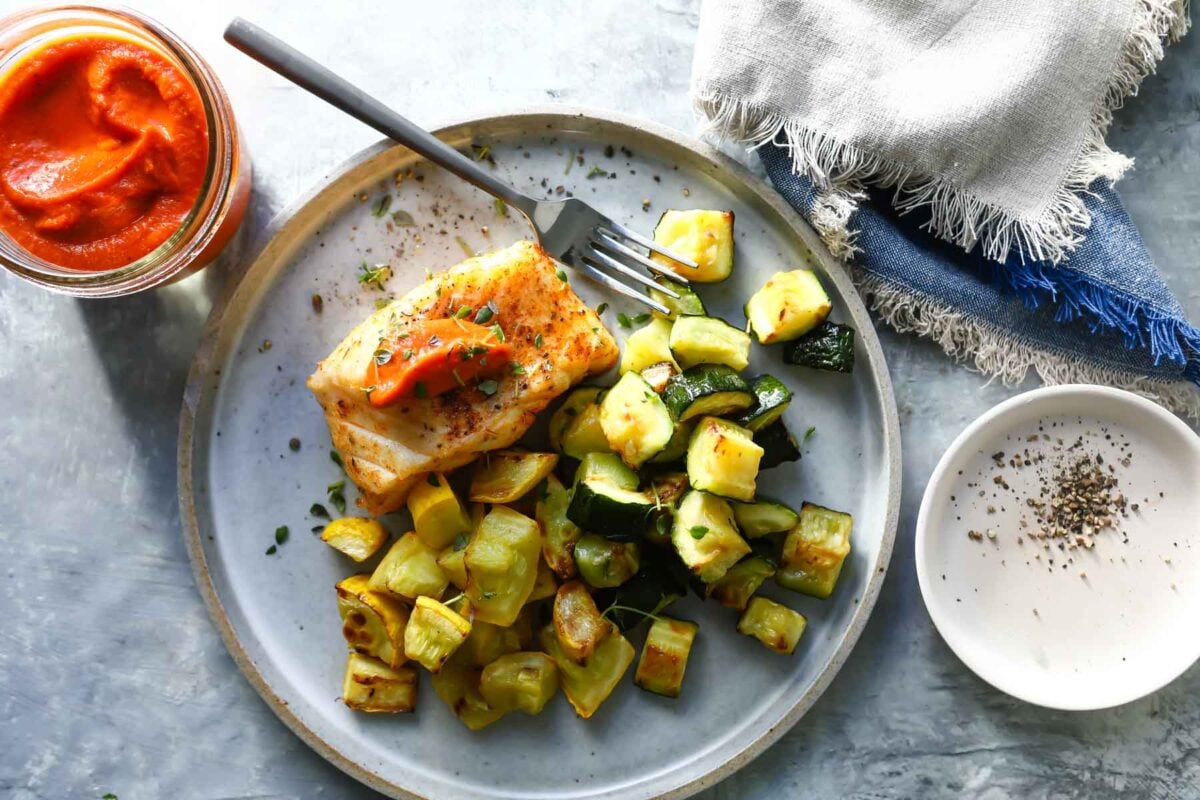 A piece of air fryer cod topped with romesco sauce served next to air fryer zucchini.
