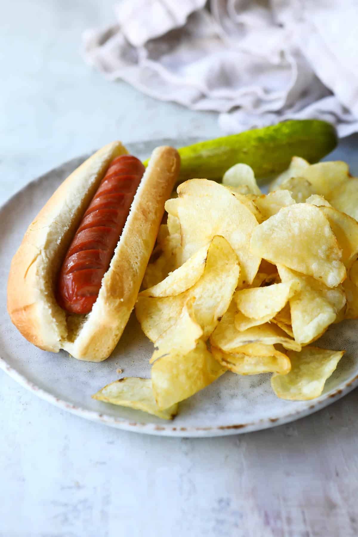 An air fried hot dog on a plate with chips and a pickle.