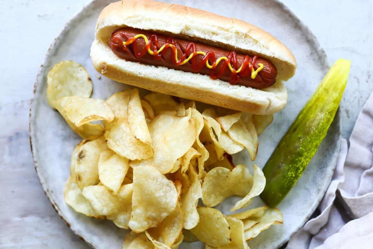 An air fried hot dog on a plate with chips and a pickle.