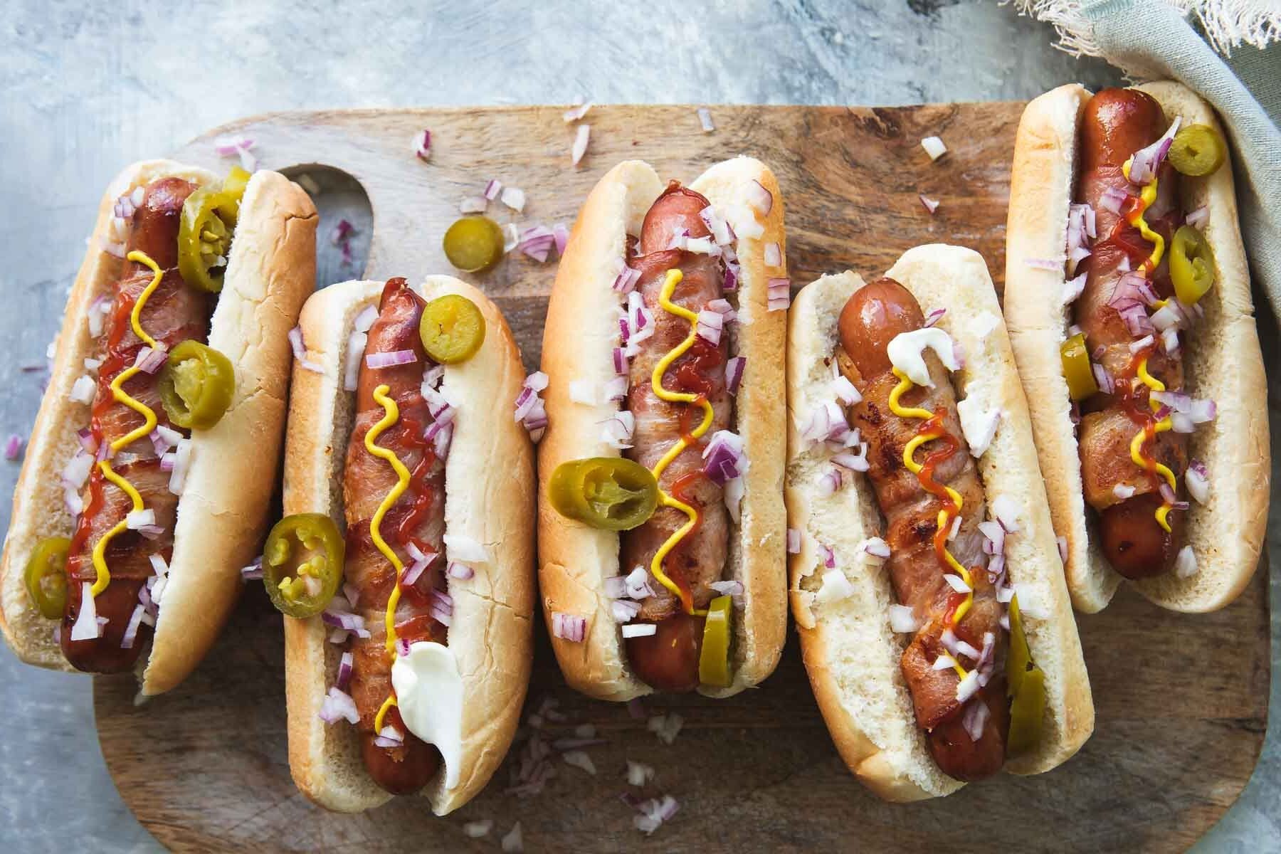 Bacon wrapped hot dogs on a wooden cutting board with toppings.