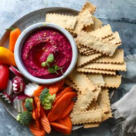 A small bowl of beet hummus surrounded by vegetables and crackers.