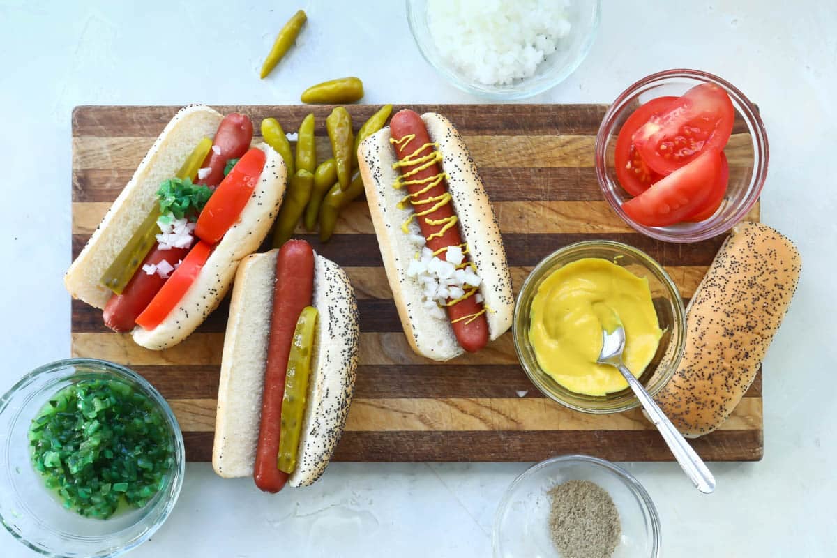 Chicago style hot dogs being assembled on a wooden cutting board.