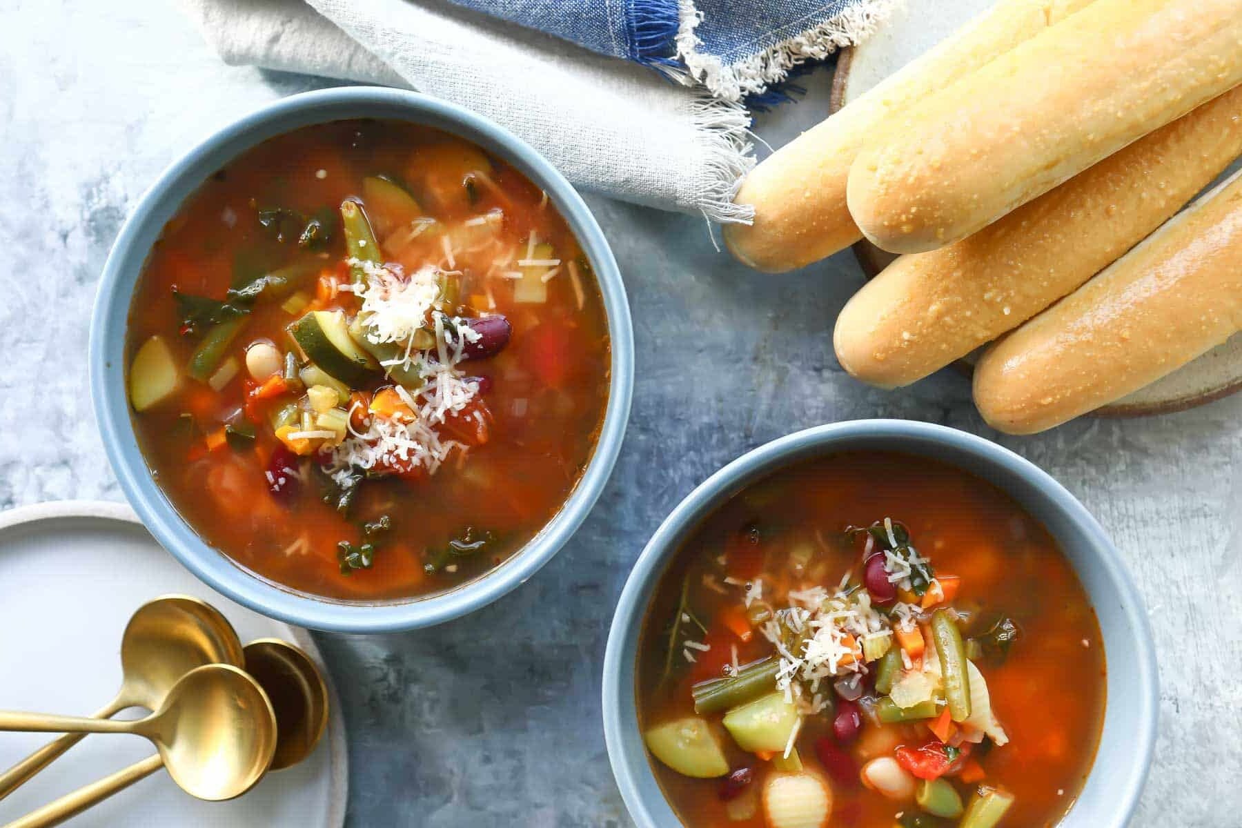 Two bowls of Olive Garden Minestrone soup next to breadsticks.
