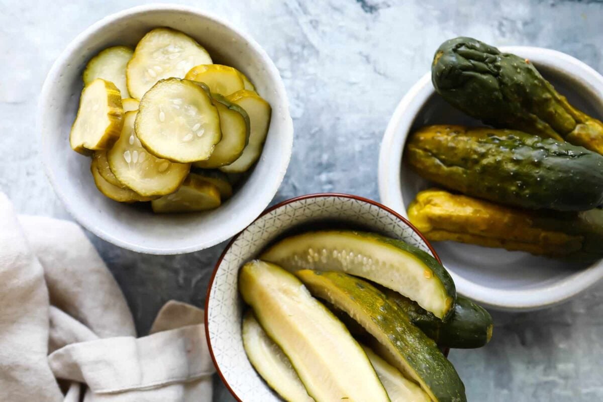 Three bowls of pickles containing pickle chips, pickle spears, and whole pickles.