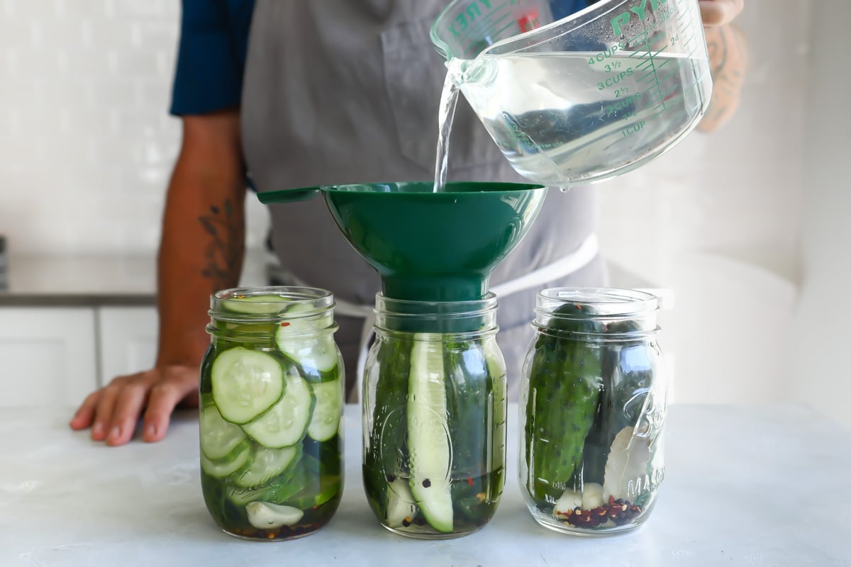 Someone adding clear liquid to mason jars of cucumbers.