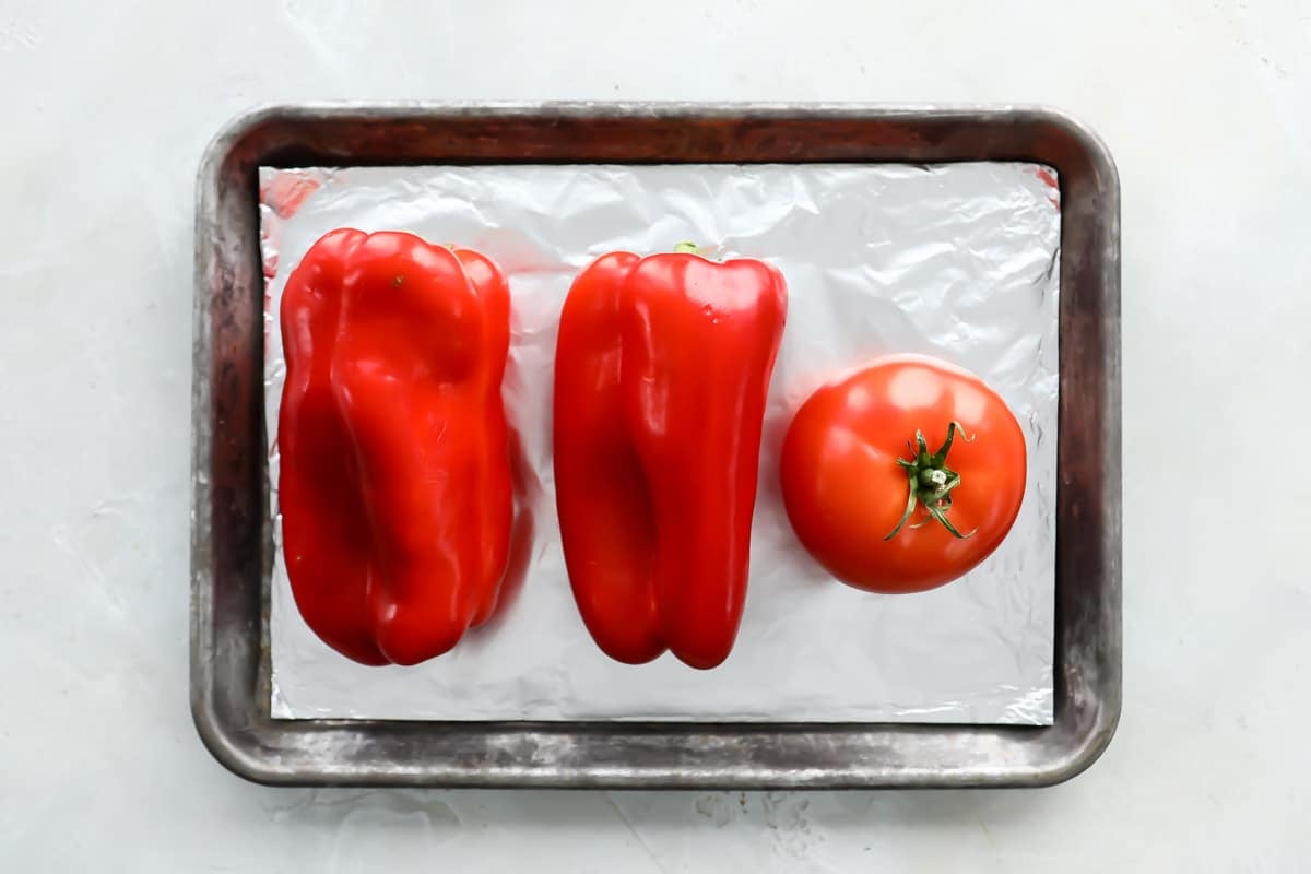 Two red peppers and a tomato on a parchment paper lined baking sheet.