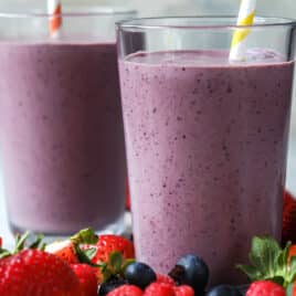 Two acai smoothies in clear glasses on a countertop surrounded by berries.