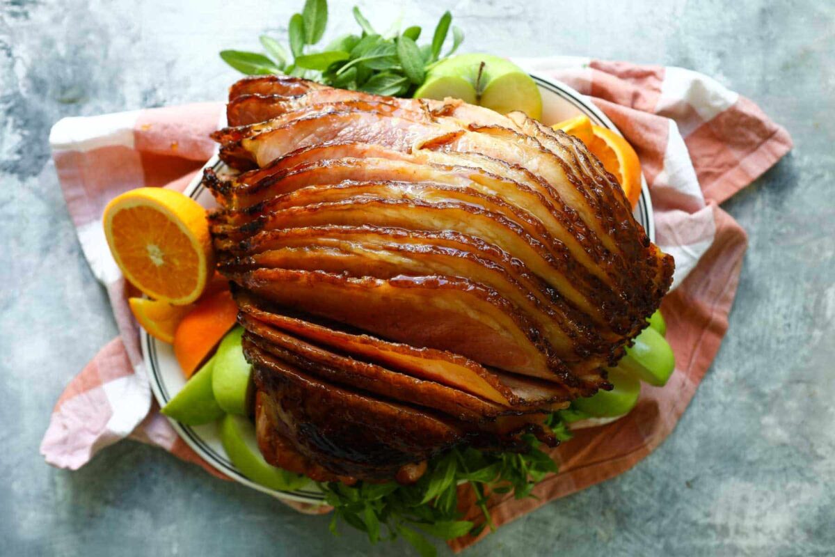 A sliced baked ham on a platter with fresh herbs and oranges.