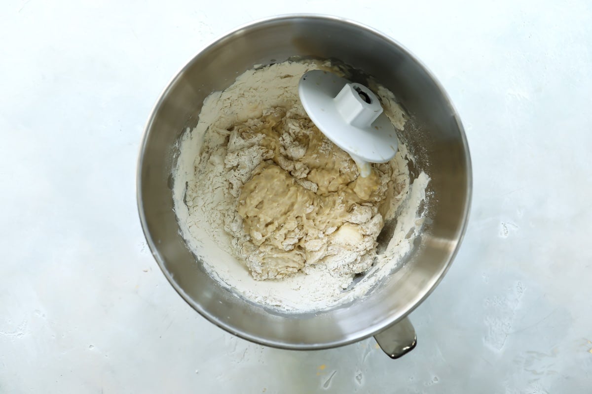 Dough and a dough hook for soft yeast dinner rolls in a silver mixing bowl.