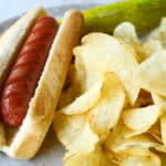 An air fried hot dog on a plate with chips and a pickle.