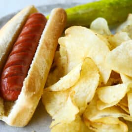 An air fried hot dog on a plate with chips and a pickle.