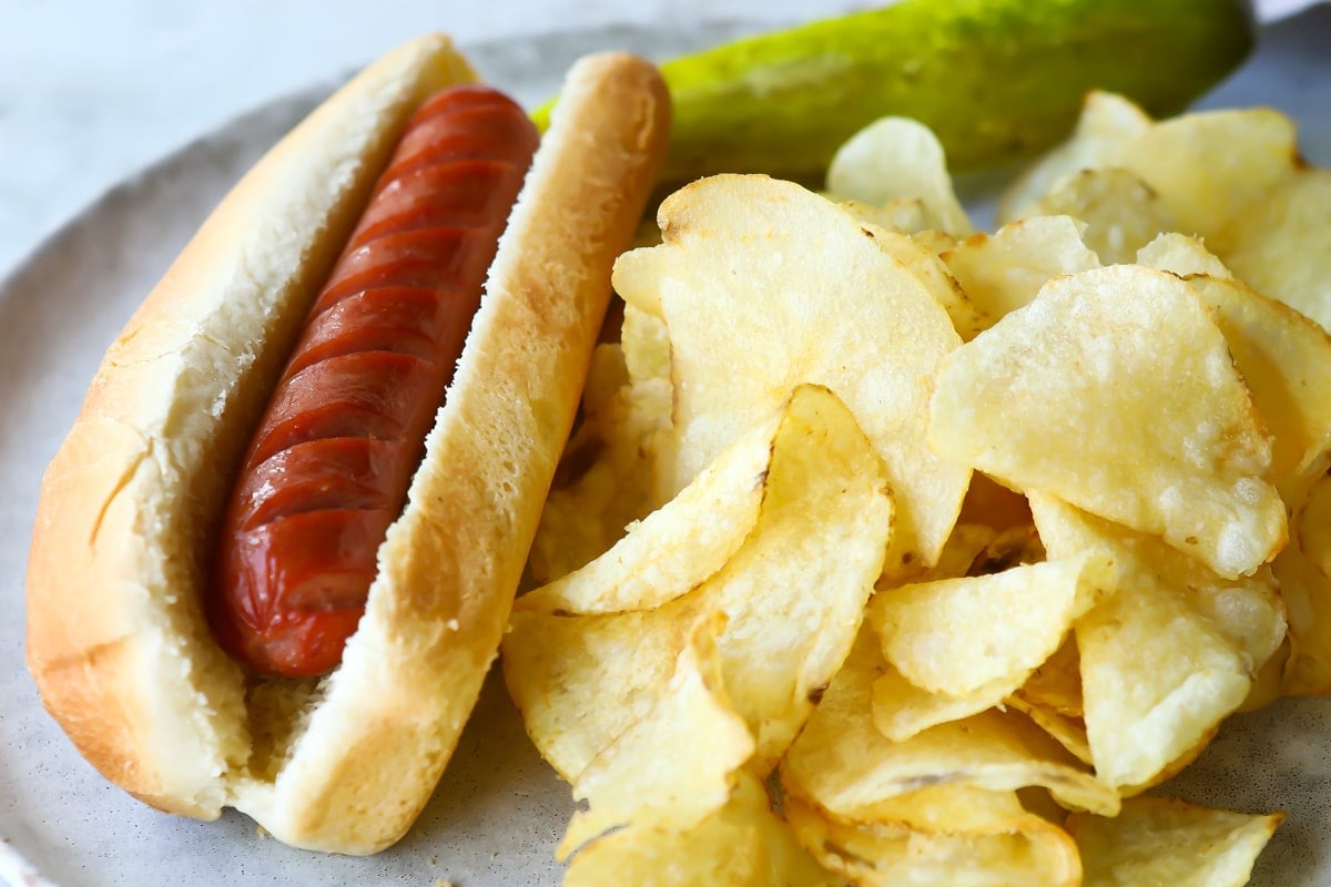 An air fried hot dog on a plate with chips and a pickle.