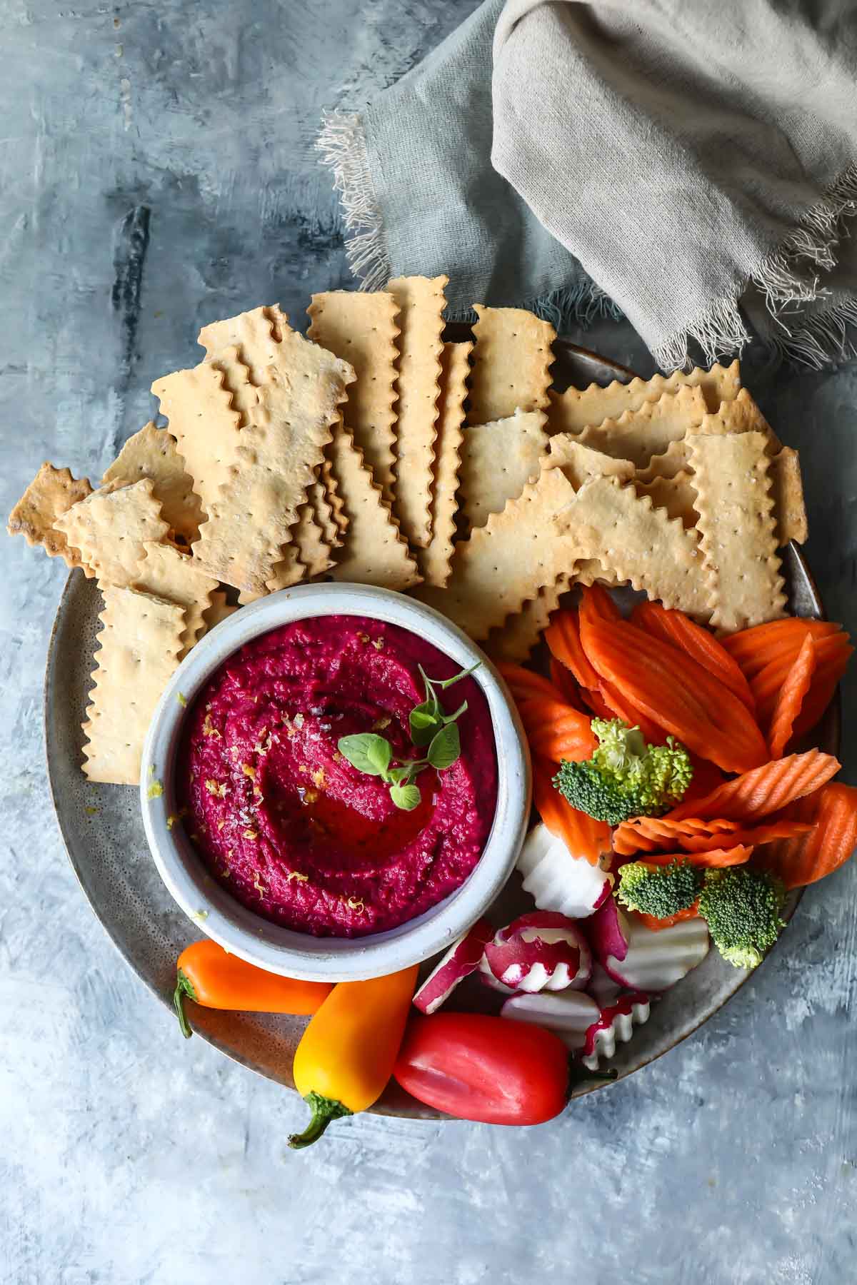 A small bowl of beet hummus surrounded by vegetables and crackers.