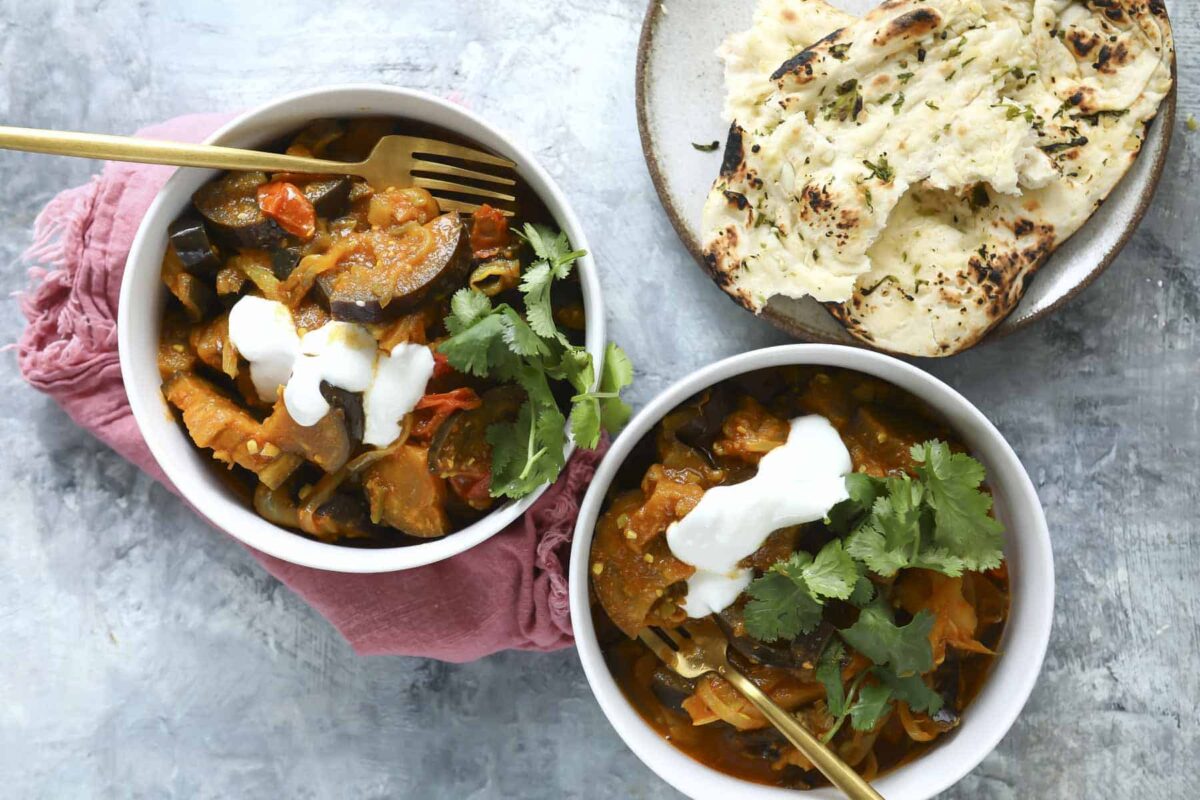 Two bowls of eggplant curry with yogurt on top and naan nearby.