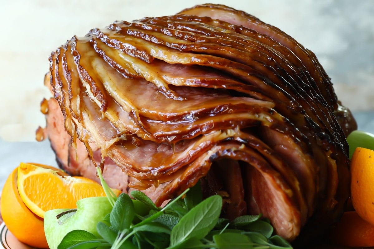 A sliced baked ham on a platter with fresh herbs and oranges.