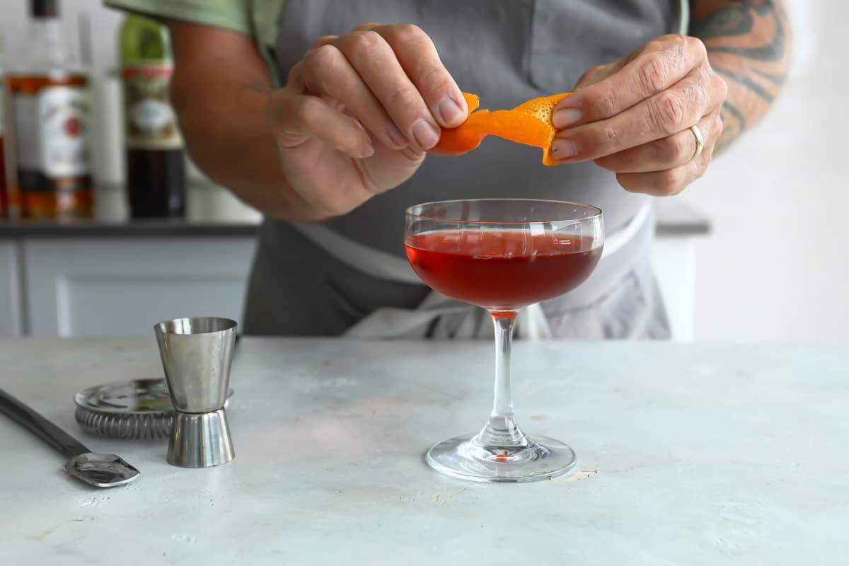 Someone twisting an orange peel to garnish a boulevardier cocktail.
