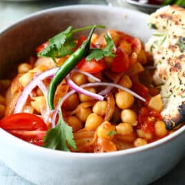 A bowl of Chana Masala with fresh veggie garnishes and naan bread on the side.