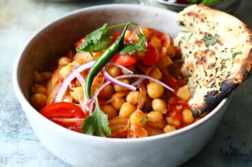 A bowl of Chana Masala with fresh veggie garnishes and naan bread on the side.