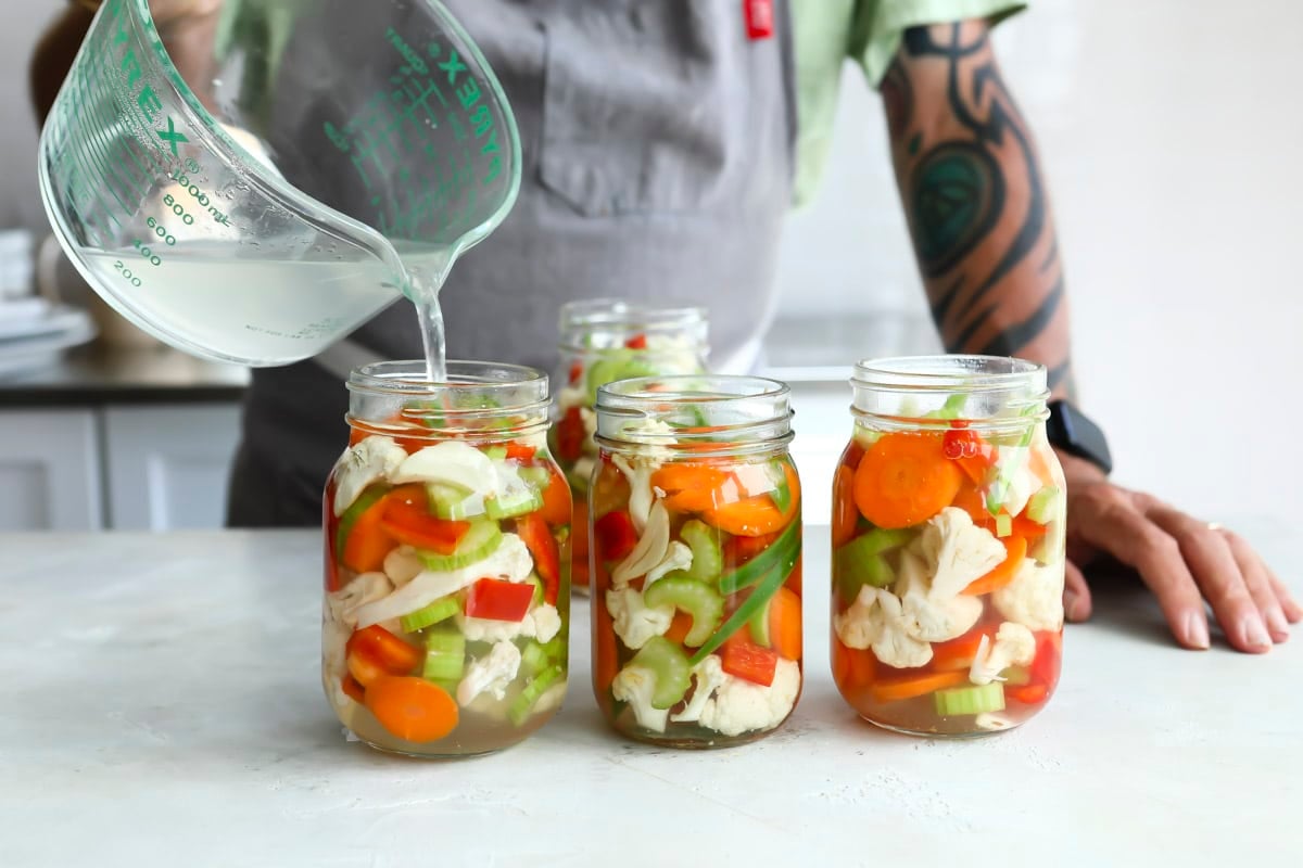 Someone pouring clear liquid into three mason jars of Giardiniera.