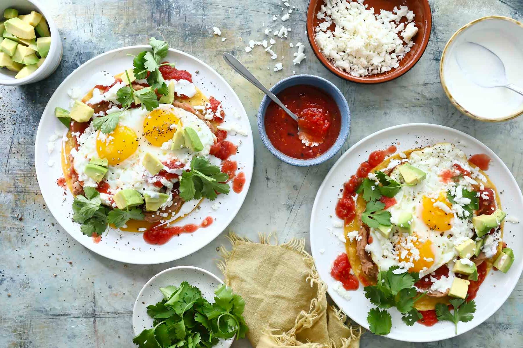 Two plates of Huevos Rancheros on a white plates surrounded by small bowls of toppings.