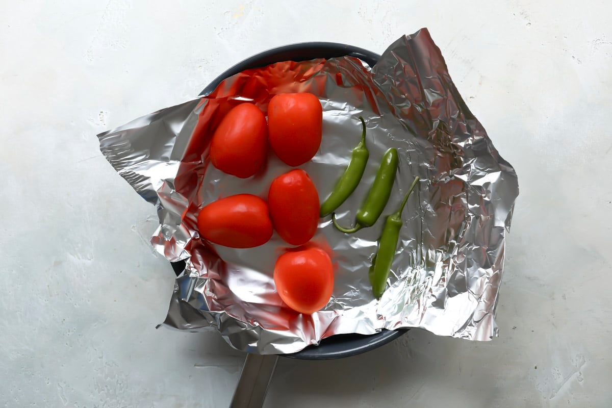 Tomatoes and chiles on aluminum foil in a skillet before being roasted.
