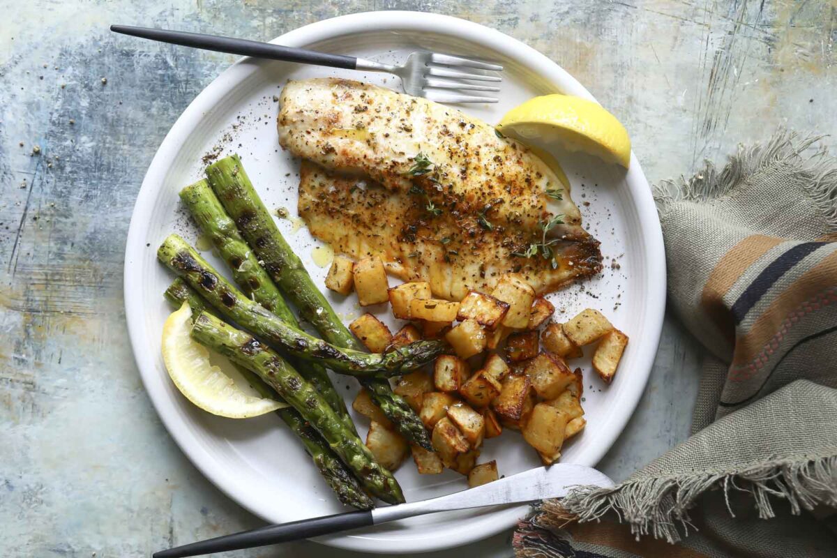 Air fryer tilapia on a plate with cubed potatoes, lemon wedges and grilled asparagus.