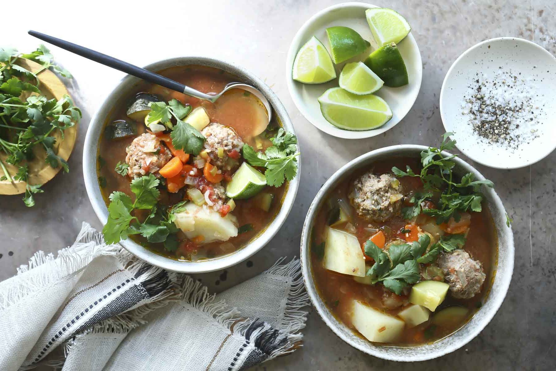 Two bowls of Albondigas soup on a countertop next to a small bowl of limes.