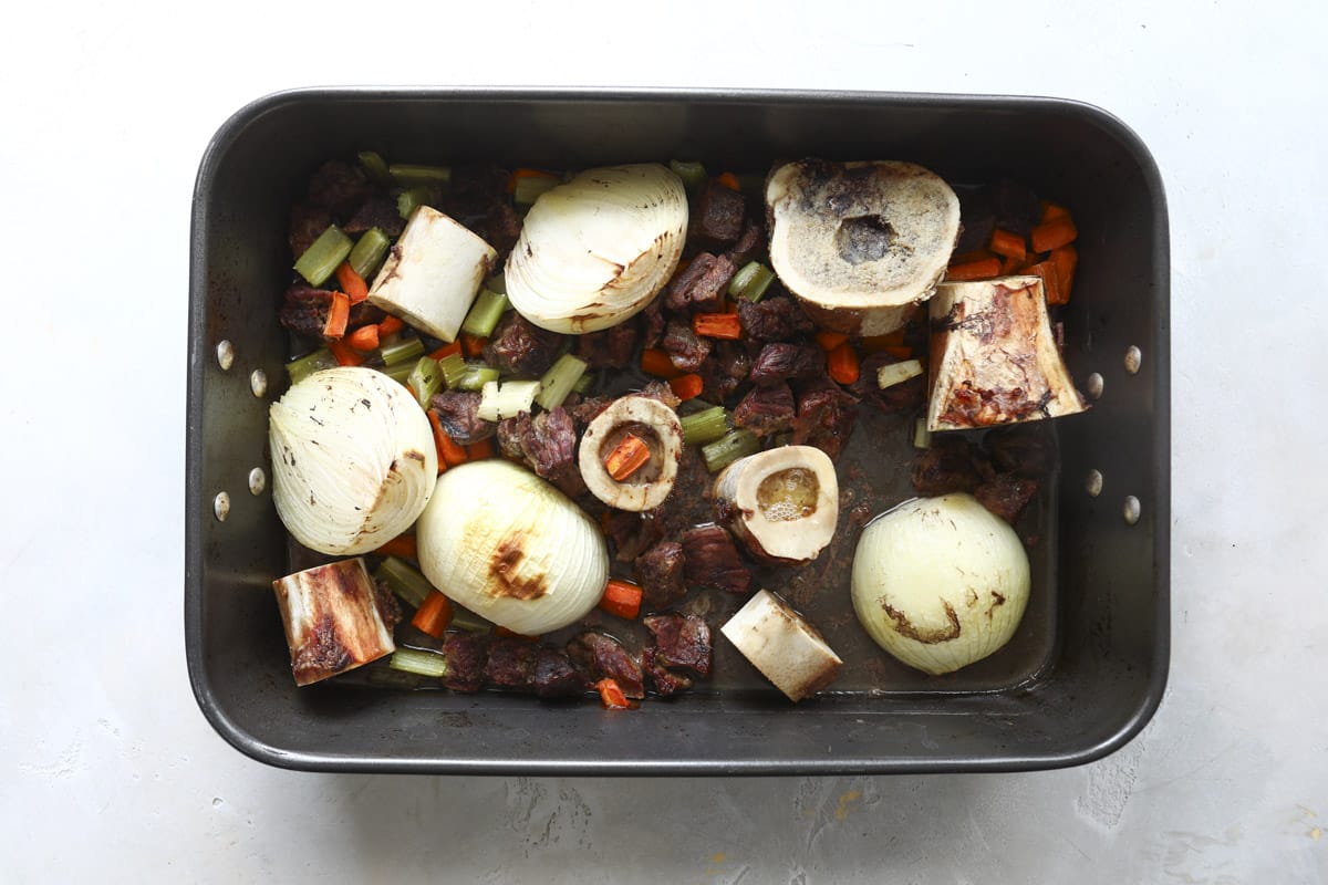 A pan of marrow bones, raw beef, and fresh vegetables after roasting.
