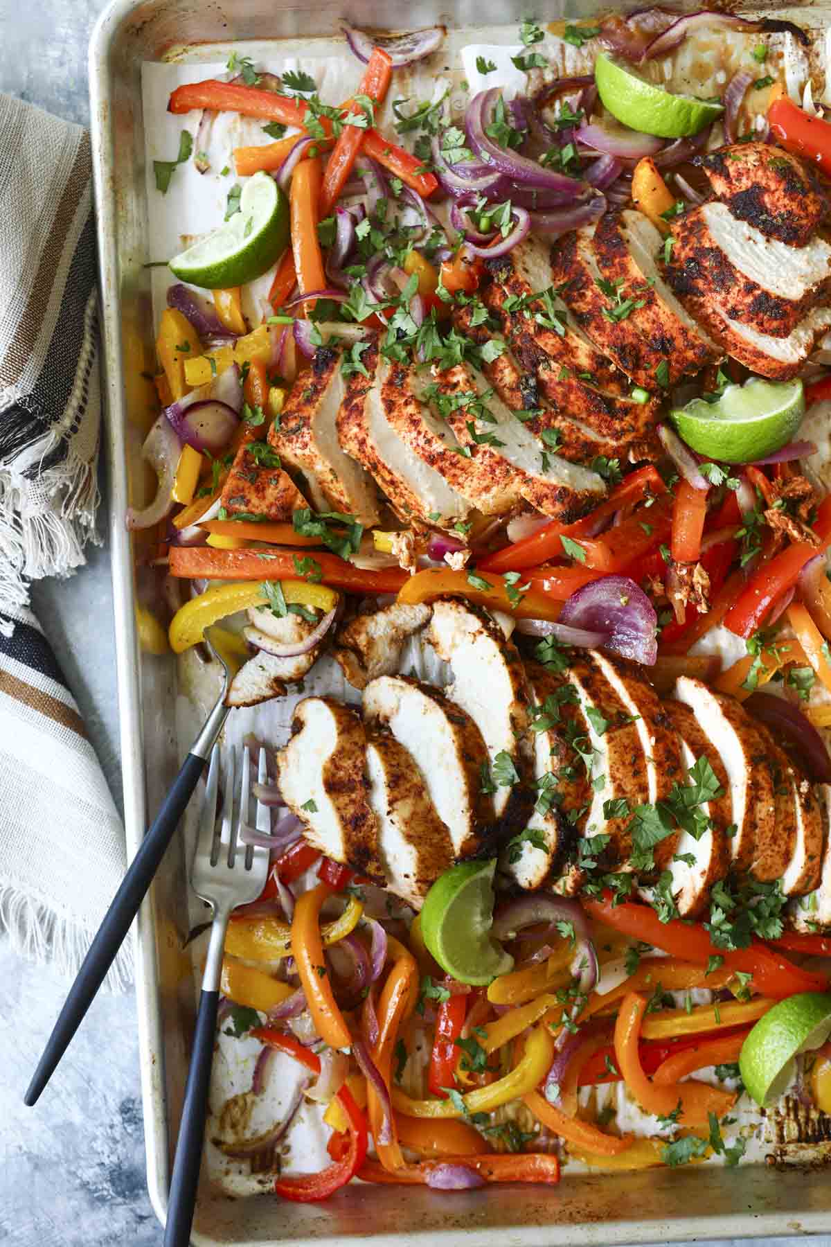 A sheet pan of chicken fajitas with sliced chicken breast, peppers, onions, and lime wedges.