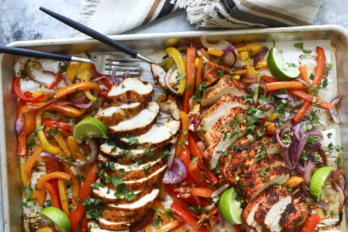 A sheet pan of chicken fajitas with sliced chicken breast, peppers, onions, and lime wedges.