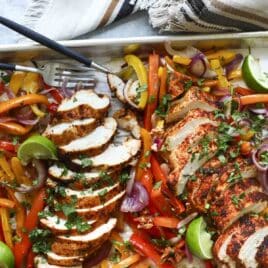 A sheet pan of chicken fajitas with sliced chicken breast, peppers, onions, and lime wedges.