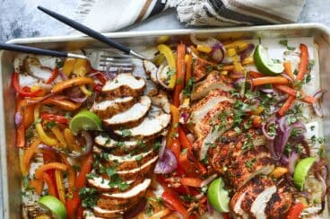 A sheet pan of chicken fajitas with sliced chicken breast, peppers, onions, and lime wedges.