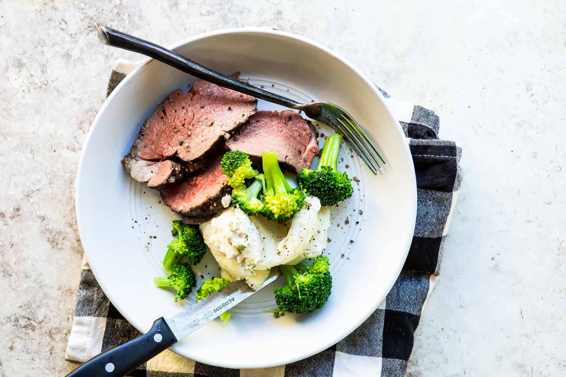 Slices of roast beef tenderloin on a plate with broccoli and mashed potatoes.