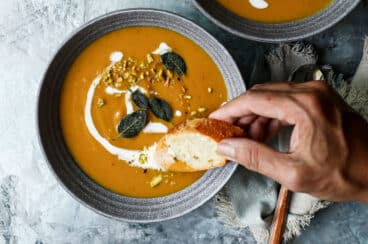 A bowl of butternut squash soup with someone dipping bread into it.