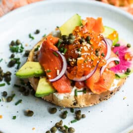 A lox bagel on a white plate with vegetables, herbs, and caviar.