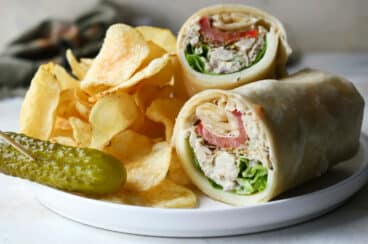 A tuna wrap on a plate with chips and a pickle.