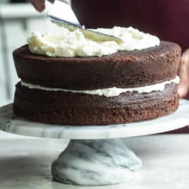 Someone applying vanilla frosting to a chocolate layer cake.